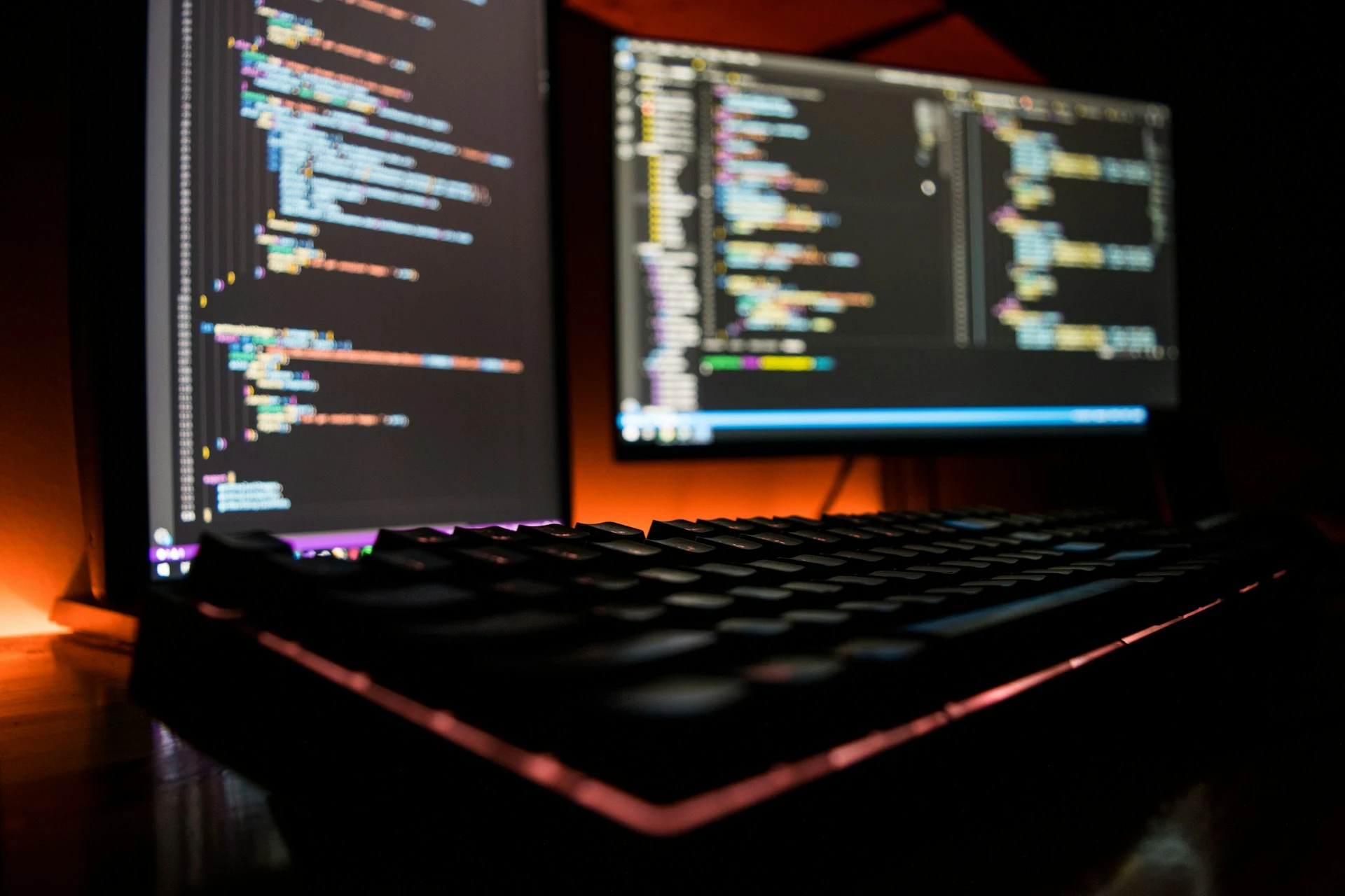 Zwei Monitore, links Vertikal positioniert, rechts Horizontal positioniert, mit Code auf den Bildschirmen zu sehen. Im Vordergrund eine Tastatur. Alles platziert auf einer Arbeitstischplatte. Hinter den Bildschirmen an der Wand und die Tastatur ist eine Orange beleuchtung zu sehen.